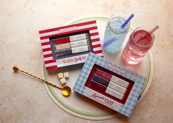 Two mason jars with colorful drinks and straws sit beside two boxes of lemonade-themed sugar cubes and a gold spoon, all placed on a round placemat.