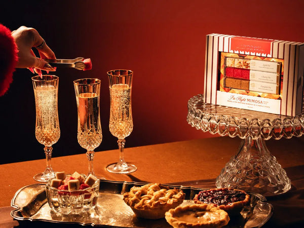 A styled food and drink scene with a hand placing a mixer cube into a champagne flute, alongside three filled flutes, pastries on a tray, and a boxed chocolate gift set labeled 'Pie Flight MImosa' on a glass pedestal against a warm red-brown background.