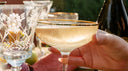 A gold-rimmed coupe glass filled with a sparkling botanical cocktail, held during an elegant outdoor Damp January social gathering