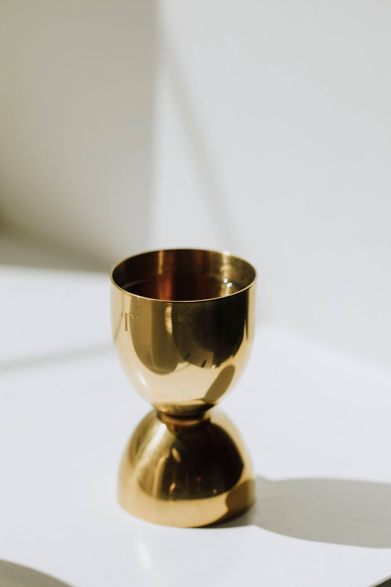 A gold-colored metal cocktail jigger with a reflective surface, positioned upright on a light-colored surface.