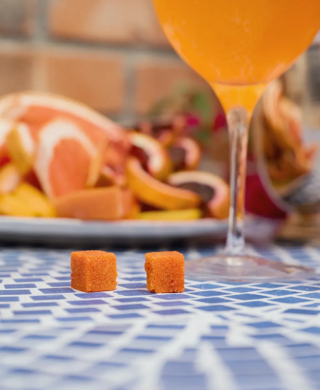 Two small orange cubes on a blue checkered tablecloth with a blurred background of a fruit orange slices and a glass with an Aperitivo spritz.