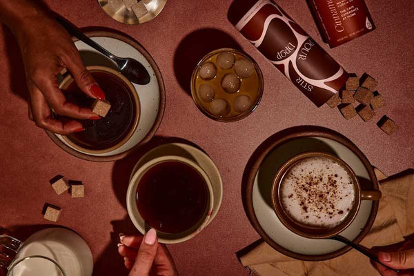 A flat lay of coffee drinks, including a cup of hot coffee, an iced drink with ice cubes, and a latte with cinnamon, alongside sugar cubes and a carton of flavored coffee.