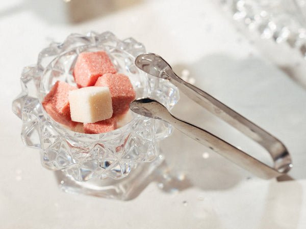 A small clear crystal glass bowl with a faceted scalloped rim holding pink and white champagne mixer cubes, with silver tongs resting beside it on a bright white surface.