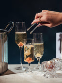 Three champagne flutes filled with sparkling wine on a table, alongside a metal ice bucket, a hand holding mini silver tongs, and a small glass bowl with champagne mixer cubes, set against a dark background.