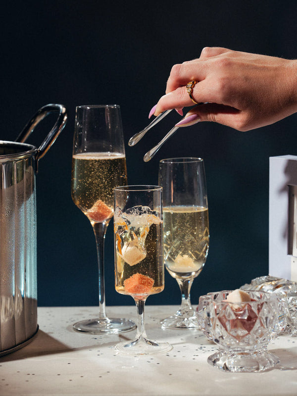 Three champagne flutes filled with sparkling wine on a table, alongside a metal ice bucket, a hand holding mini silver tongs, and a small glass bowl with champagne mixer cubes, set against a dark background.