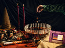 A styled Halloween-themed scene with a large ribbed glass punch bowl filled with dark red drink, surrounded by candles, metallic cups,cocktail mixer cubes, a witch hat, and a package labeled 'Witches Brew Punch' on a draped table against a dark curtain. A hand in a green sleeve drops cubes into the bowl.