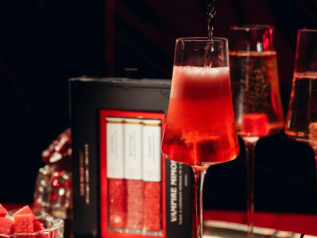 A close-up of a red mimosa in a stemmed glass with a dark liquid being poured into it, set against a dark background with a black and red product box labeled 'VAMPIRE mimosa' and other glasses in the scene.