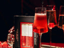 A close-up of a red mimosa in a stemmed glass with a dark liquid being poured into it, set against a dark background with a black and red product box labeled 'VAMPIRE mimosa' and other glasses in the scene.