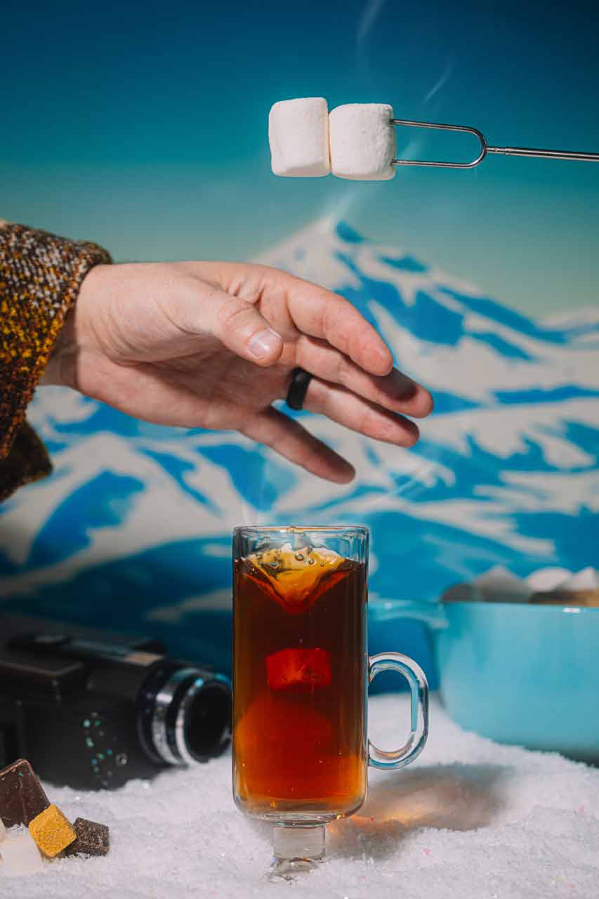 Hand holding a glass of hot beverage with marshmallows over a snowy mountain background