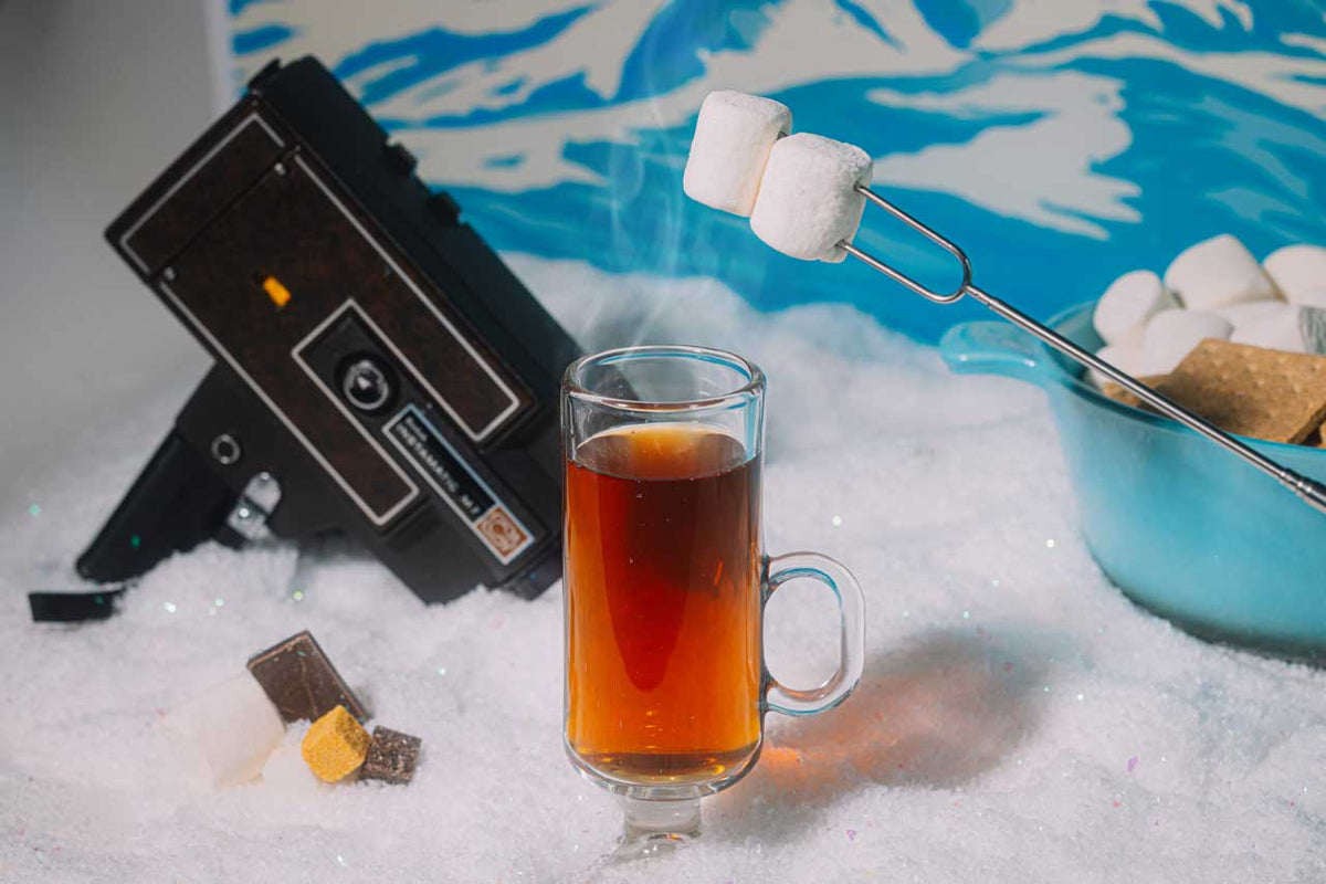 Glass of amber liquid with marshmallows on a skewer, chocolate pieces, and a vintage camera in a snowy setting.
