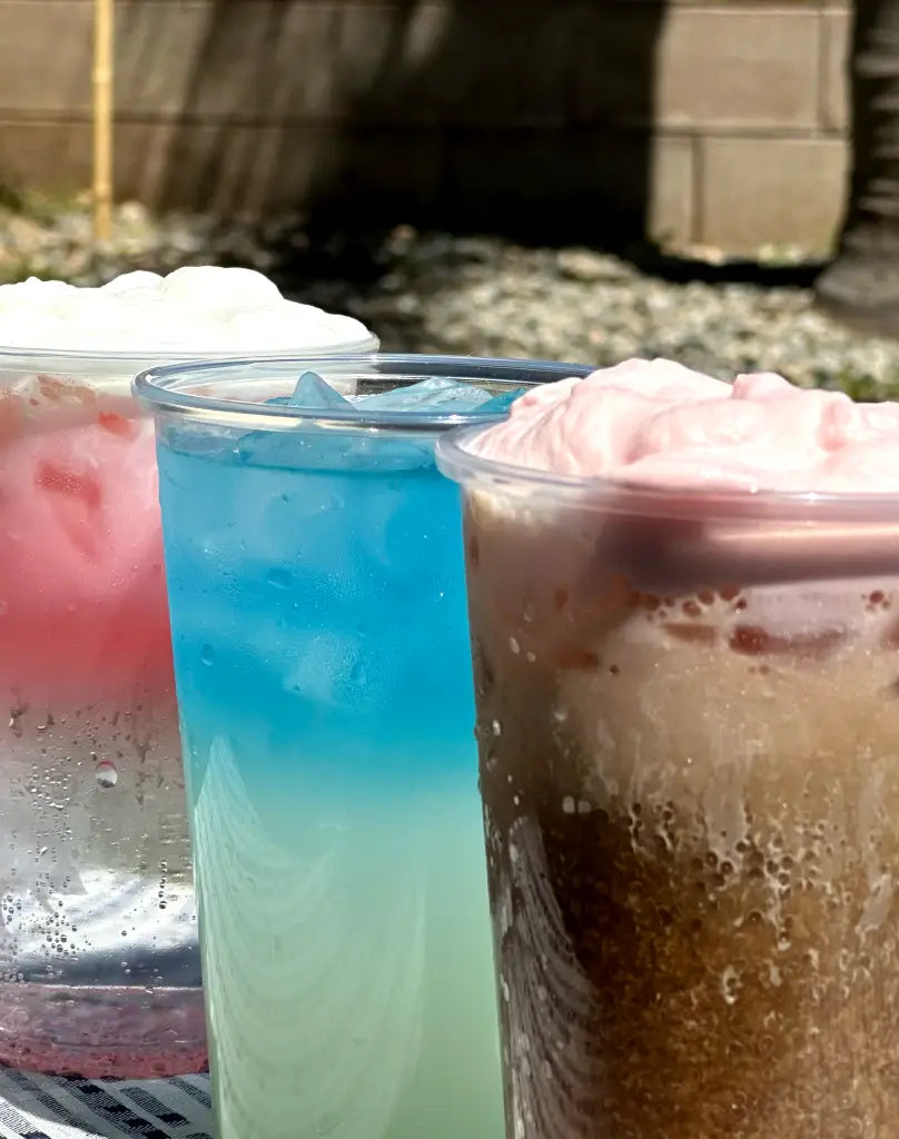 Three colorful drinks in clear cups with whipped cream on top, featuring pink, blue, and brown beverages, sitting on a surface with light coming from above.