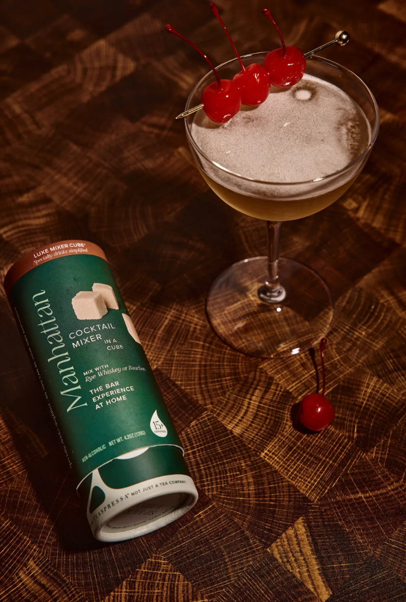 Manhattan cocktail mixer can next to a cocktail with cherries on a wooden surface