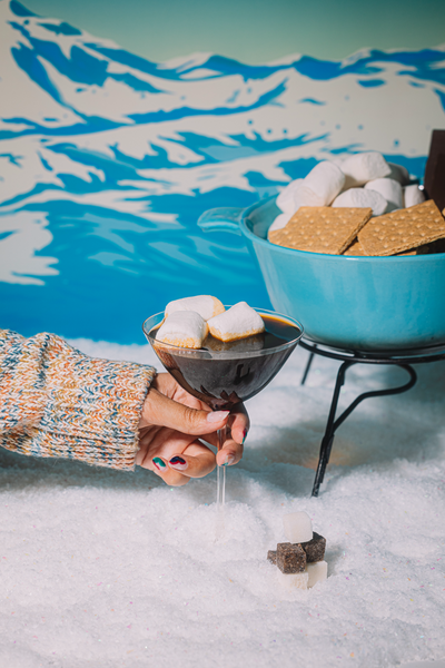 Hand holding a cocktail with marshmallows in front of a s'mores setup against a mountainous background