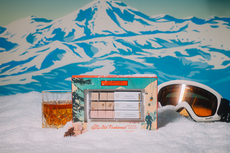 Snowy mountain landscape with a box of Chai Eggnog Old Fashioned cocktails, a glass of cocktail, and goggles.