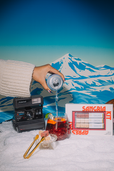 Hand pouring a La croix into a winter sangria. With a mountain and vintage polaroid camera in the background