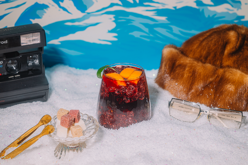 Glass of Winter Sangria drink with orange slices, a fur hat, glasses, and gold mini tongs on a snowy surface with blue and white patterned background.