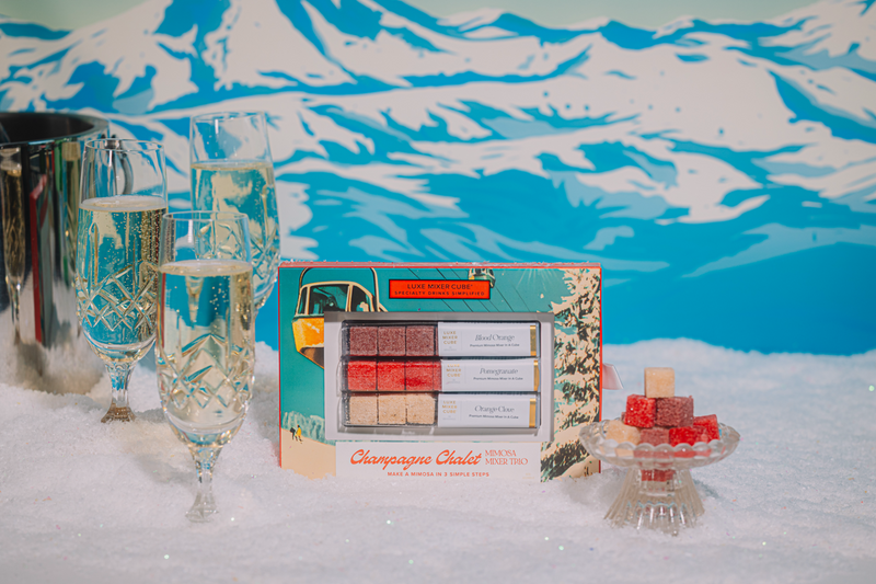 Champagne glasses with a Champagne Chalet Mixer trio gift box and small tray of Luxe Mixer Cubes on a snowy background