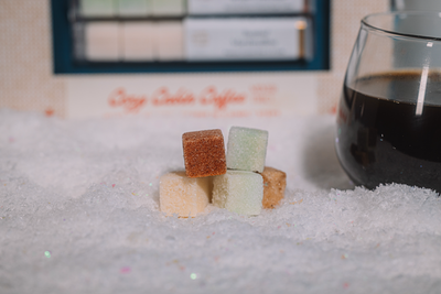 brown, light green and cream Luxe Mixer cubes stacked on a snowy surface with a glass of coffee in the background.