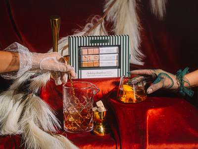 Person with decorative gloves holding a glass of amber liquid next to a small box on a red surface.