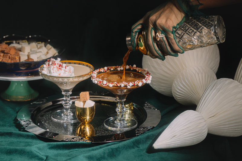 Person pouring a drink into a cocktail glass on a dark surface with decorative elements.