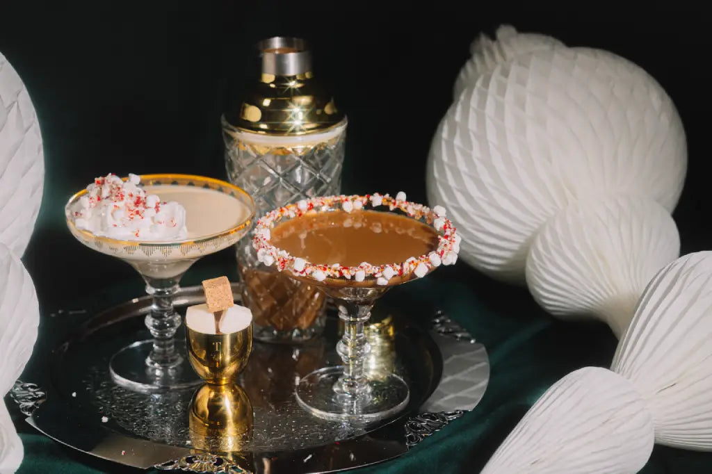 Two cocktail glasses with a brown liquid and decorative rim on a dark surface with white spherical objects in the background.