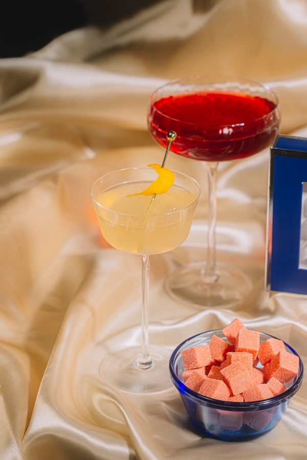 Two cocktails on a satin surface with a small bowl of pink cubes.