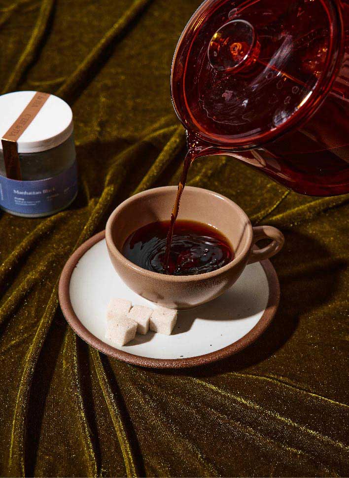 A cup of coffee is being poured from a glass pot, with sugar cubes on a plate beside it, set against a textured green fabric background.