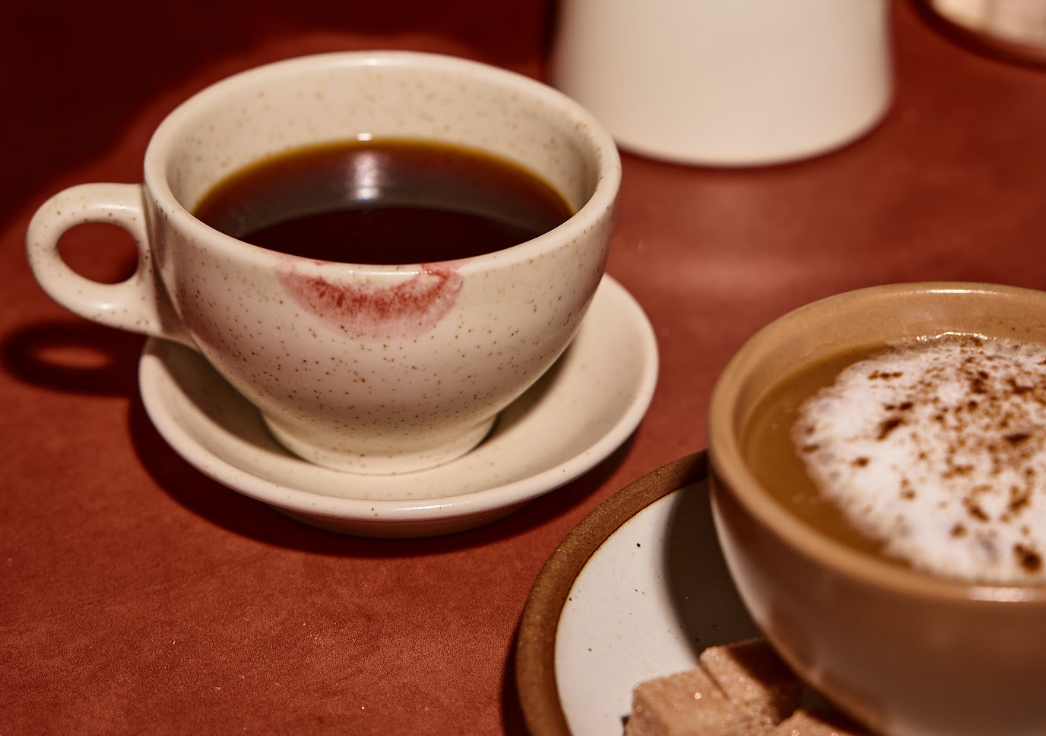 A coffee cup with a distinct lip mark, accompanied by a smaller cup of cappuccino and a plate with sugar cubes, set on a warm-colored surface.