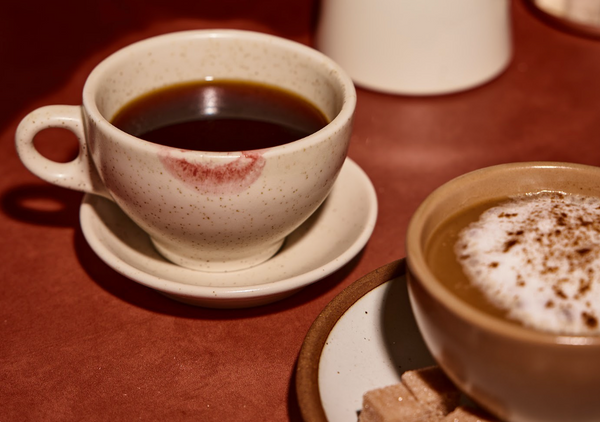 A coffee cup with a distinct lip mark, accompanied by a smaller cup of cappuccino and a plate with sugar cubes, set on a warm-colored surface.