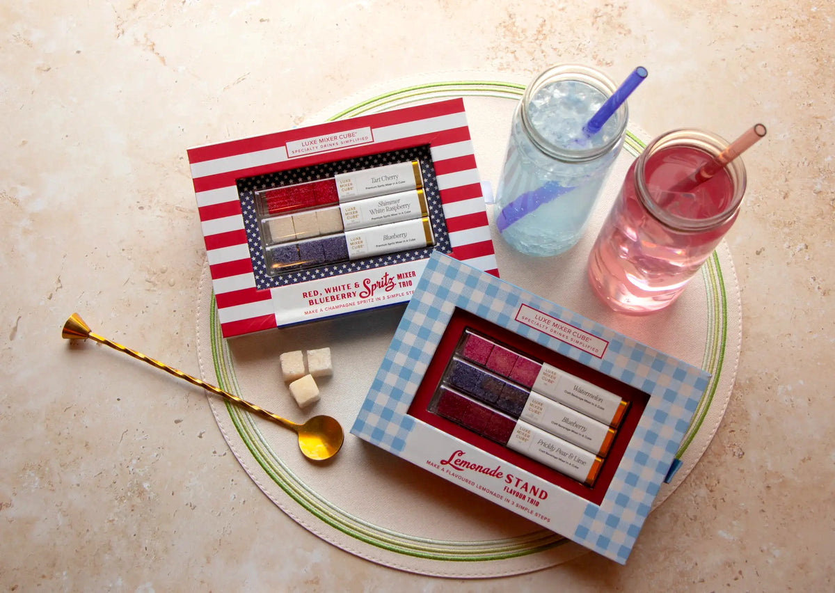 Two mason jars with colorful drinks and straws sit beside two boxes of lemonade-themed sugar cubes and a gold spoon, all placed on a round placemat.