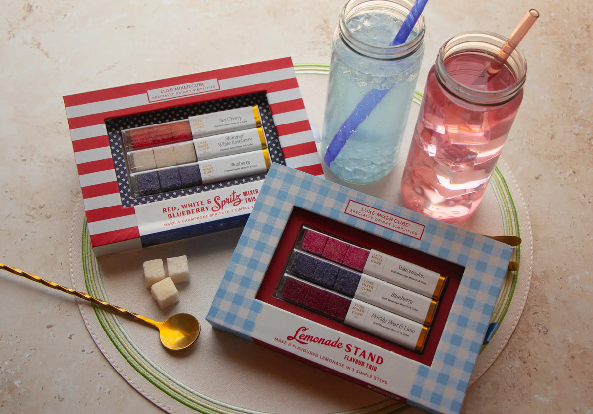 Two mason jars with colorful drinks and straws sit beside two boxes of lemonade-themed sugar cubes and a gold spoon, all placed on a round placemat.