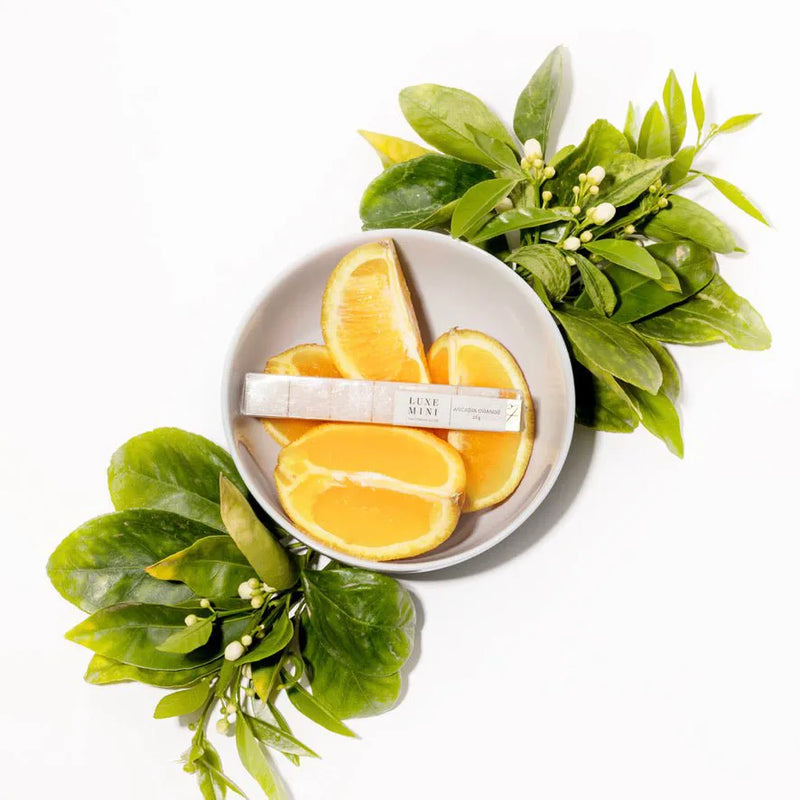 A small grey bowl containing orange slices, accompanied by fresh green leaves and a labeled product tube resting on top of the fruit.