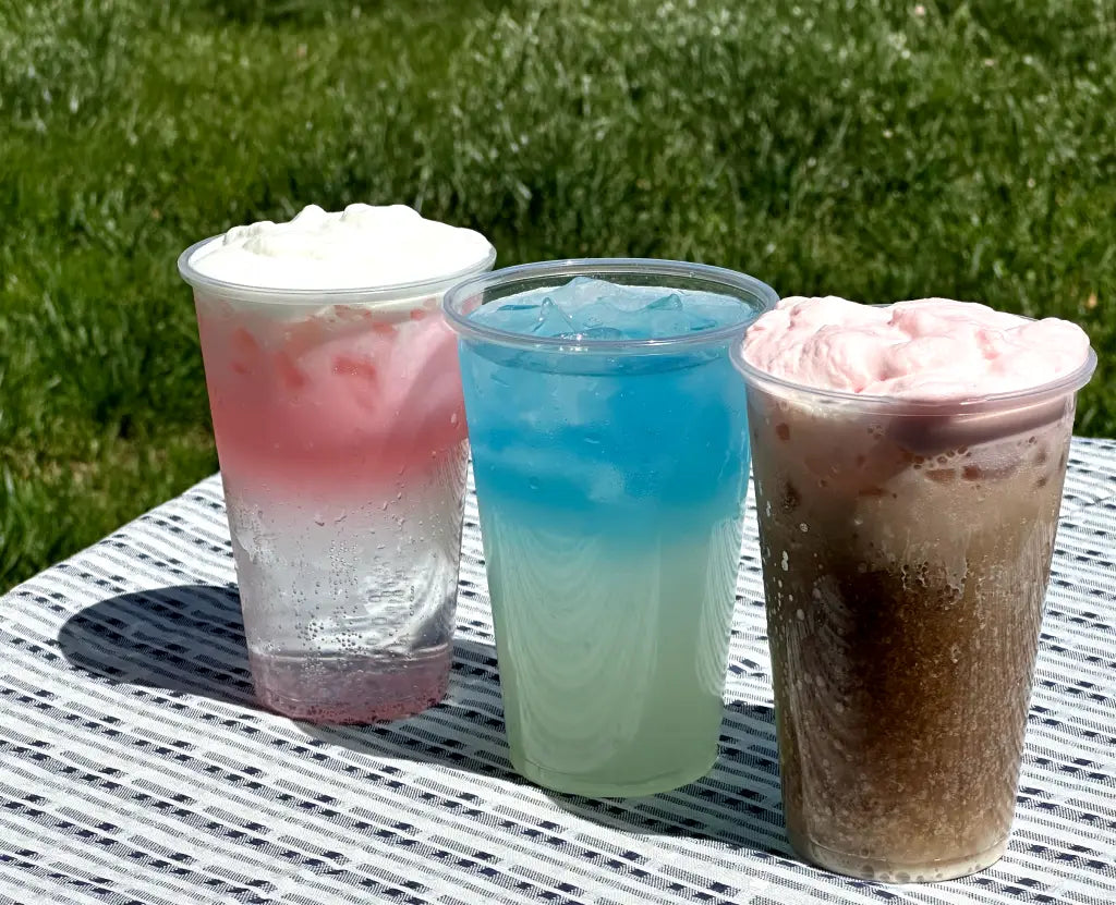 Three colorful drinks in clear cups on a table: a pink layered drink with whipped cream, a blue icy drink, and a brown fizzy drink topped with pink foam.