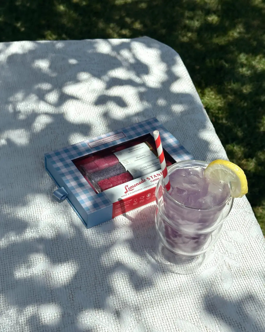 A glass of purple lemonade with ice, a red-striped straw, and a lemon slice sits on a white tablecloth next to a box of candies. Dappled sunlight and leafy shadows cover the table; grass is visible in the background.