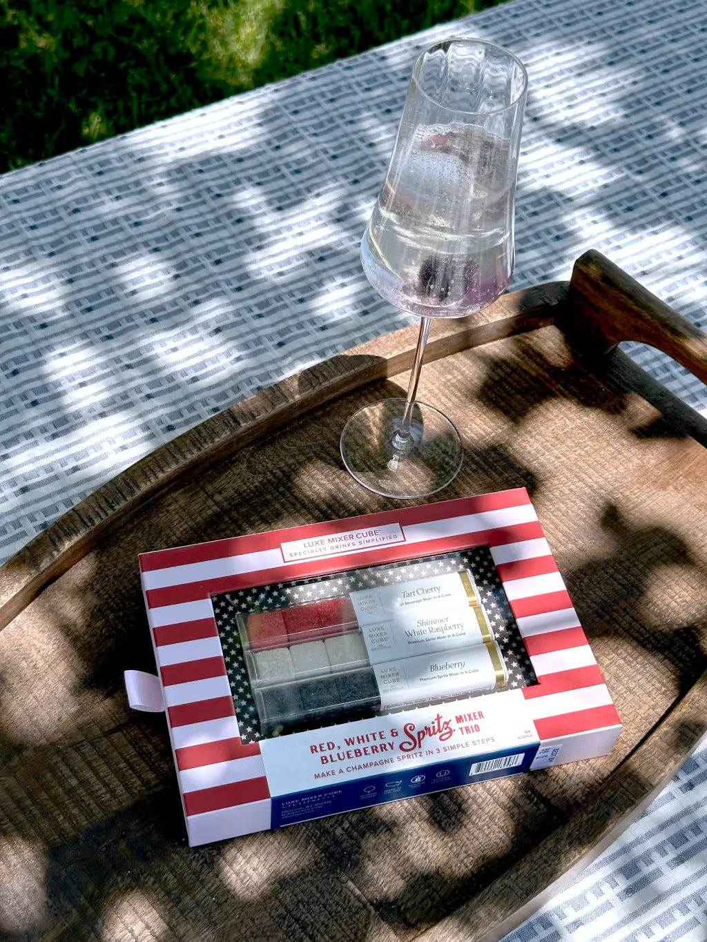 A glass of sparkling drink and a patriotic Spritz kit with red, white, and blue packaging sit on a wooden tray atop a patterned outdoor tablecloth in dappled sunlight.