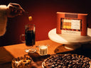 Hand adding sugar cube to a tall glass latte. Pecan Pie Latte Mix Kit, whole pecan pie, and milk on a table with warm, cozy lighting.