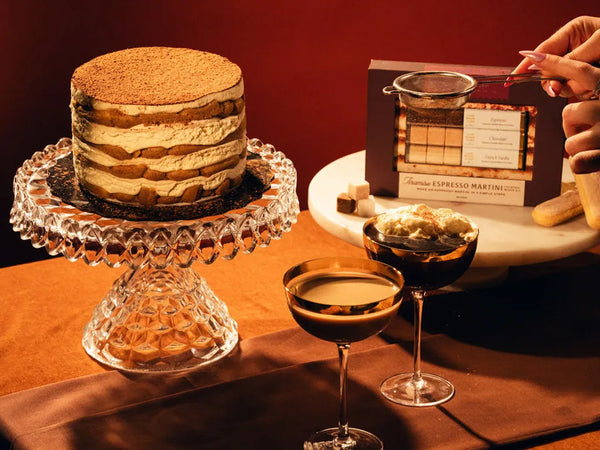 A layered tiramisu cake on a crystal cake stand, accompanied by two espresso martini cocktails, with a box labeled "Tiramisu Espresso Martini" in the background.