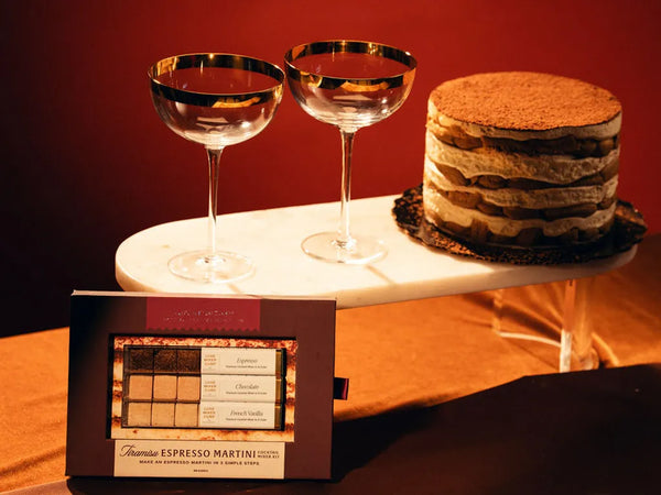 Two elegant coupe glasses with gold rims, a layered tiramisu cake, and a box labeled "Tiramisu Espresso Martini Cocktail Maker Kit" are arranged on a marble surface with a warm background.