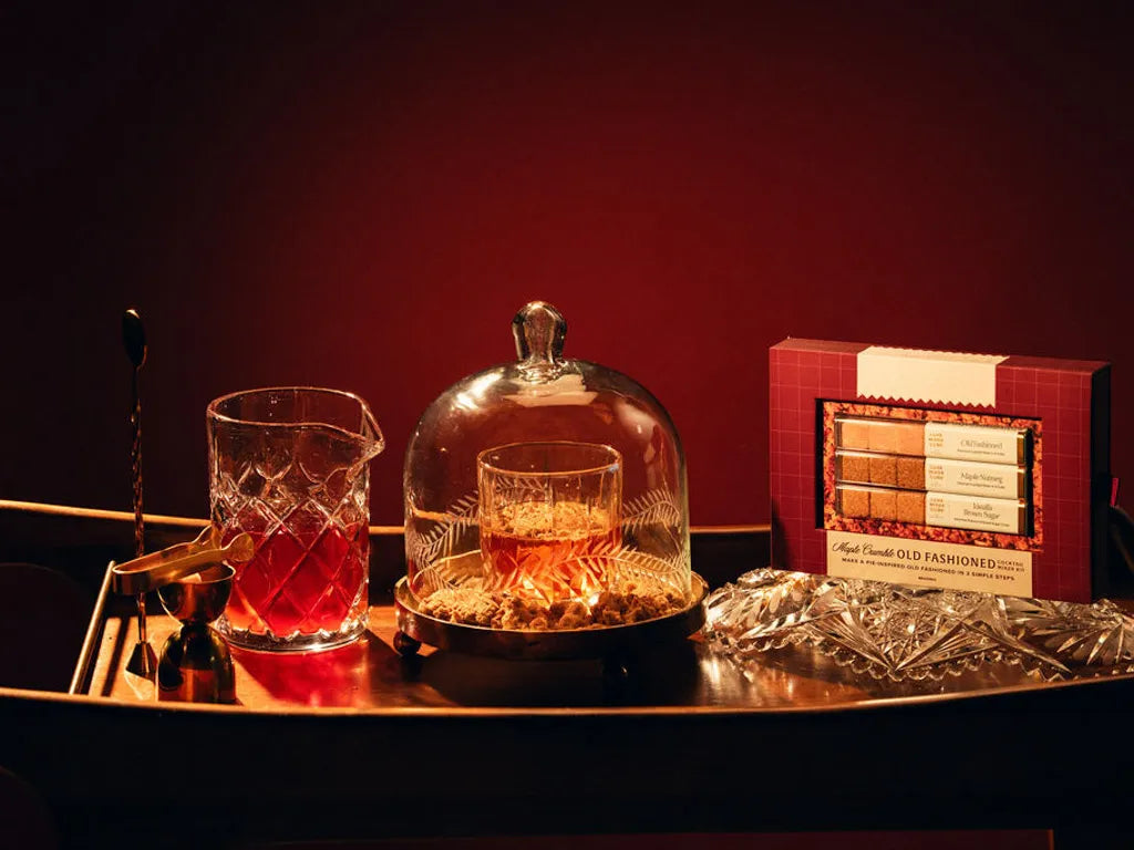 A wooden tray with a crystal glass containing an Old Fashioned cocktail, a glass jug, a covered dish with snacks, a gold jigger, and a boxed cocktail mixer set, all against a dark background.