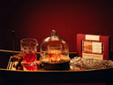 A wooden tray with a crystal glass containing an Old Fashioned cocktail, a glass jug, a covered dish with snacks, a gold jigger, and a boxed cocktail mixer set, all against a dark background.
