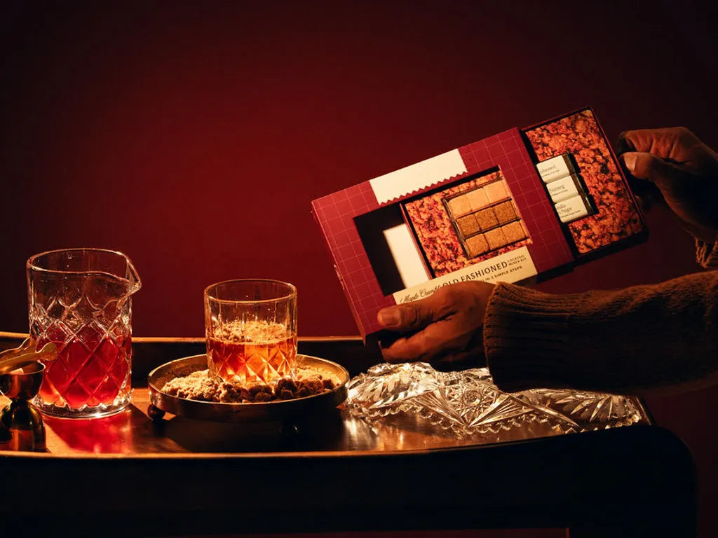 A wooden tray with a crystal glass containing an Old Fashioned cocktail, Hands holding up Gift set, next to a glass jug, a covered dish with snacks, a gold jigger, and a boxed cocktail mixer set, all against a dark background.