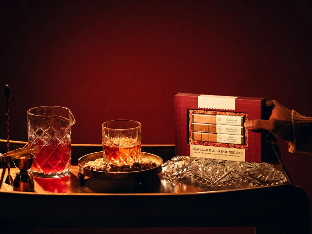 A wooden tray with a crystal glass containing an Old Fashioned cocktail, Hands holding up Gift set, next to a glass jug, a covered dish with snacks, a gold jigger, and a boxed cocktail mixer set, all against a dark background.