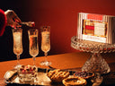 A styled food and drink scene with a hand placing a mixer cube into a champagne flute, alongside three filled flutes, pastries on a tray, and a boxed chocolate gift set labeled 'Pie Flight MImosa' on a glass pedestal against a warm red-brown background.