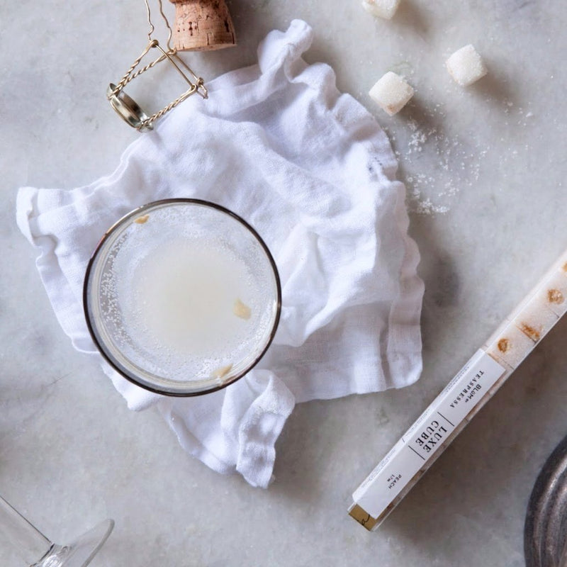 A top-down view of a glass with a light-colored beverage, placed on a white cloth. Nearby are a cork, peach infused sugar cubes, and a drink stirrer.
