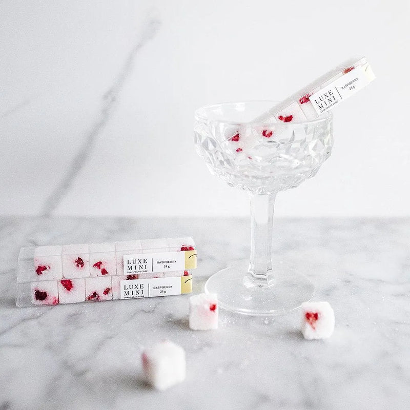 A crystal glass filled with white cubes that have raspberry pieces, accompanied by two packs labeled "Luxe Mini Raspberry" on a marble surface.