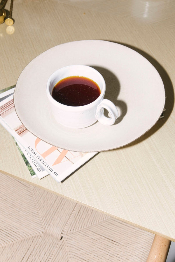 White cup of coffee on a white plate with a magazine underneath on a wooden table.