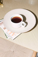White cup of coffee on a white plate with a magazine underneath on a wooden table.