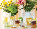 A collection of colorful cocktails, lemonades, and arnold palmers garnished with fruits, arranged on a table with vibrant yellow flowers in the background.