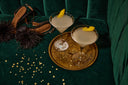 Two cocktails in elegant glasses, garnished with lemon twists, are placed on a round gold tray. Nearby, stylish high-heeled shoes with fluffy decorations rest on a green velvet surface, accompanied by scattered gold sequins.